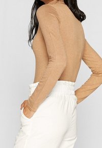Top de manga larga y ajustado en material de punto color beige con un diseño de espalda recta. Combinado con pantalones de cintura elástica blanca de tiro alto.