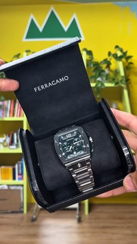 Ferragamo rokas pulkstenis melnā dāvanu kastē. Pulkstenim zaļa ciparnīca, sudraba metāla siksniņa, kvadrātveida korpuss un gaismu atstarojošas atzīmes.
