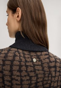 Strickpullover mit einem geometrischen braun-schwarzen Muster, hohem Rippkragen und einem silbernen Emblem auf der Rückseite. Weicher Stoff.
