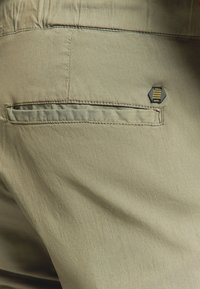 Khaki broek met een gladde textuur, met een achterzak en een zwarte hexagonale tag met gele strepen.