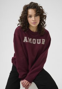 Sweatshirt borgogna con la scritta "AMOUR" in lettere di crema testurizzate. Presenta maniche lunghe e una vestibilità rilassata. Indossato con pantaloni neri.