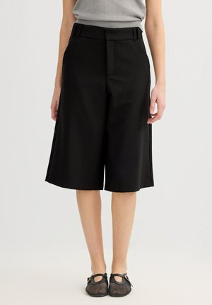 Mujer con pantalones cortos negros de pierna ancha hasta la rodilla, top sin mangas gris claro y zapatos Mary Jane de malla negra, de pie sobre un fondo liso.