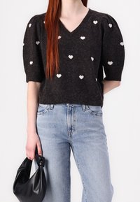 Pull en maille gris foncé avec motifs de cœurs blancs, col en V, manches courtes bouffantes. Associé à un jean taille haute bleu clair et un sac noir.