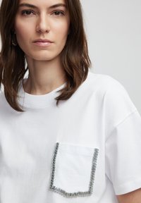 Witte T-shirt met korte mouwen, voorzien van een gestructureerde zak met zilveren versieringen langs de randen. Zacht katoenen stof; relaxte pasvorm.
