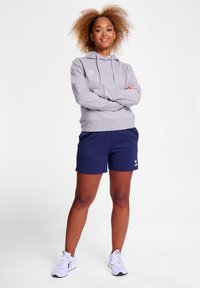 Grauer Pullover-Hoodie mit einer Fronttasche, kombiniert mit dunkelblauen Shorts. Modell trägt weiße Sportschuhe, Arme verschränkt, lässige Haltung.