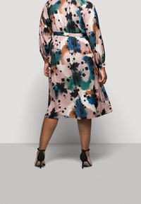 Multicolored bloemenjurk in lichtroze, met lange mouwen, een cinched taille en een vloeiende rok; gecombineerd met zwarte gympen met hoge hakken.
