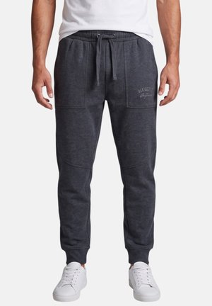 Hombre vistiendo pantalones jogger gris oscuro con bolsillos delanteros y cintura con cordón, combinados con zapatillas blancas y una camiseta blanca.