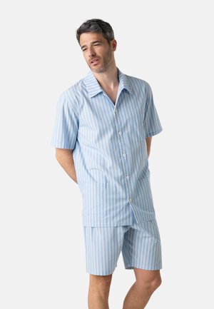 Homme portant une chemise à manches courtes bleu clair boutonnée et un short assorti avec des rayures verticales blanches et beiges, debout les mains derrière le dos.