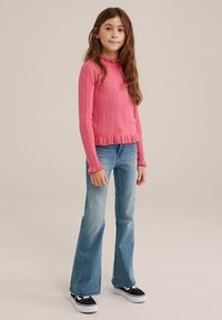 Růžový žebrovaný svetr s volánovým výstřihem, kombinovaný se světlomodrými zvonovými džínami a černými teniskami s bílou podrážkou, na neutrálním pozadí.