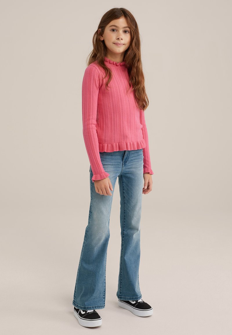 Růžový žebrovaný svetr s volánovým výstřihem, kombinovaný se světlomodrými zvonovými džínami a černými teniskami s bílou podrážkou, na neutrálním pozadí.