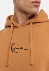 Brun hoodie med dragsnören, med en svart broderad signatur. Slät textur, avslappnad passform och inga synliga mönster eller detaljer.