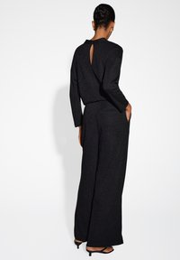 Completino nero con una vestibilità rilassata, maniche lunghe e dettaglio sul retro a forma di chiave. Tessuto testurizzato con una leggera brillantezza. Caratterizzato da gambe larghe e tasche.