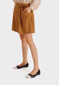 Pantaloni corti di cotone marroni con vita regolabile, tasche laterali e vestibilità rilassata, abbinati a ballerine nere in vernice con punta color crema.