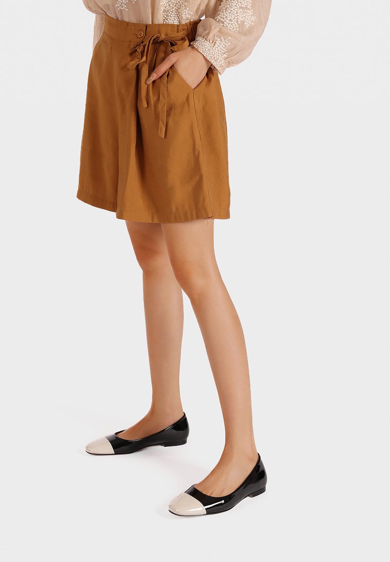 Pantaloni corti di cotone marroni con vita regolabile, tasche laterali e vestibilità rilassata, abbinati a ballerine nere in vernice con punta color crema.