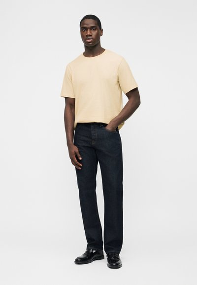 Άνδρας που στέκεται με το χέρι στην τσέπη, φορώντας μια μπεζ κοντομάνικη μπλούζα, σκούρα τζιν και μαύρα παπούτσια, μπροστά σε απλό λευκό φόντο.