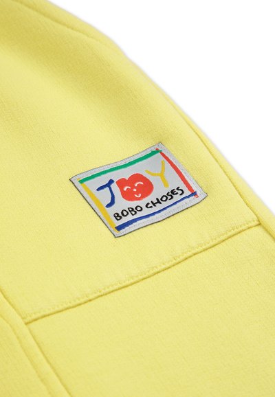 El tejido del sudadera amarilla presenta un parche cuadrado colorido con el texto "ALEGRÍA" y una cara sonriente, enmarcado por rayas coloridas y el nombre de la marca en la parte inferior.