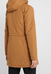 Brązowa kurtka o długości do połowy uda z kapturem. Wykonana z gładkiego, wodoodpornego materiału, ma dopasowaną talię oraz boczne kieszenie.