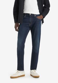 Vaqueros de mezclilla de un azul oscuro con un diseño de pierna recta, que presentan bolsillos frontales y detalles de costura sutiles, llevados con zapatillas blancas.