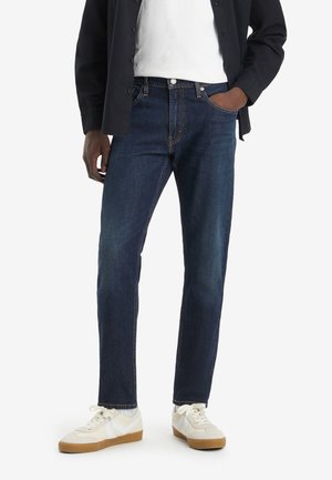 Dark blue denim jeans with a straight leg design, featuring front pockets and subtle stitching details, worn with white sneakers.