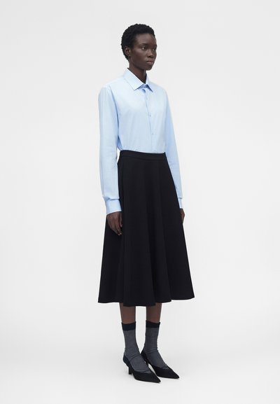 Γυναίκα στέκεται φορώντας ανοιχτό μπλε πουκάμισο με κουμπιά, μαύρη midi φούστα, γκρι κάλτσες και μαύρα μυτερά παπούτσια σε απλό γκρι φόντο.