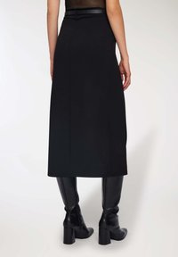 Donna che indossa una gonna midi nera con uno spacco posteriore, abbinata a stivali in pelle neri con tacco alti fino al ginocchio, in piedi su uno sfondo bianco.