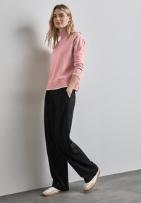 Hellrosa Pullover mit hohem Kragen und geripptem Saum, kombiniert mit schwarzen weiten Hosen. Das Model steht auf einem grauen Boden und trägt weiße Sneaker.