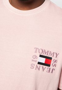 Jasnoróżowa bawełniana koszulka z małym haftowanym logo "TOMMY JEANS 85" oraz prostokątnym emblematem w kolorze czerwonym, białym i czarnym.