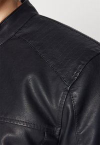 Giacca di pelle nera con dettagli trapuntati sulle spalle, texture liscia e colletto aderente. Cuciture visibili lungo le cuciture.
