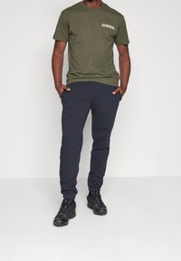 Olijfgroene t-shirt met korte mouwen en een borstzak, gecombineerd met donkerblauwe joggerbroeken. Zwarte sportieve schoenen met textuuroverzetten. Eenvoudige, casual outfit.