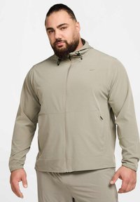 Lichtgrijze Nike-jas met rits en capuchon, voorzien van voorzakken, verstelbare trekkoorden en een gladde, lichtgewicht stof.