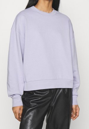 Osoba ubrana w jasnofioletową, oversizeową bluzę z długimi rękawami oraz czarne spodnie z eko skóry na jednolitym tle.