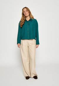 Tyrkysová košeľa na gombíky s textúrovaným povrchom, skombinovaná s voľnými béžovými nohavicami a tmavo hnedými šľapkami, čím vytvára uvoľnený outfit.