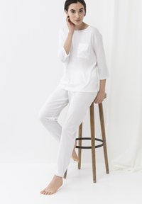 Witte katoen loungewear set met een langärmelige top met een satijnen zak en lichte broek met elastische boorden, tegen een neutrale achtergrond.