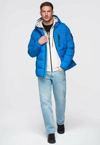 Sininen toppatakki hupulla, sivutaskuilla ja vetoketjullisella rintataskulla. Päällä valkoinen huppari, yhdistetty vaaleisiin farkkuihin ja mustiin kenkiin.