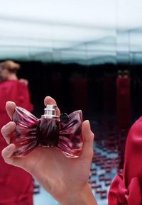Un flacon de parfum en forme de nœud, avec une couleur rose clair et un rouge foncé, doté d'un spray argenté et d'un bouchon noir, tenu dans une main. Arrière-plan réfléchissant.