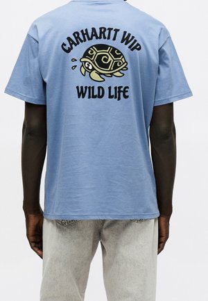 Person iført en lyseblå t-shirt med en tegneserie-skildpadde og teksten "Carhartt WIP Wild Life" trykt på ryggen.