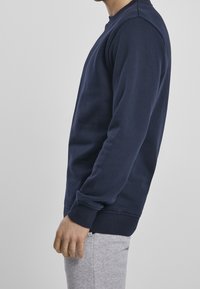 Felpa blu navy in misto cotone, collo a giro, polsini e orlo a costine, con vestibilità regolare; indossata con pantaloni della tuta grigi.