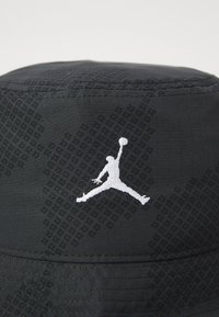 Zwarte bucket hat met een gladde textuur, voorzien van een geborduurd wit Jordan-logo aan de voorkant en subtiele patroondetails overal.