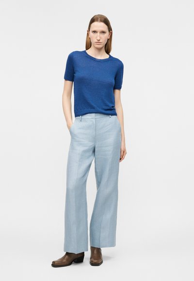 Γυναίκα που στέκεται με τα χέρια στις τσέπες ενός ανοιχτού μπλε παντελονιού φαρδιάς γραμμής, φορώντας μπλε πουλόβερ με κοντά μανίκια και καφέ μπότες αστραγάλου.