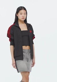 Chaqueta deportiva negra con rayas rojas, combinada con un top corto negro y una falda de mezclilla gris. La chaqueta cuenta con una cremallera y un logotipo.