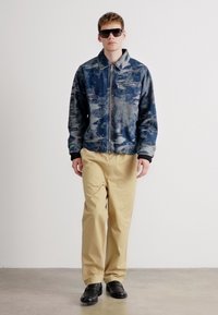Chaqueta azul con estampado y cremallera, pantalones beiges y zapatos negros. El modelo lleva gafas de sol. Textura suave, corte casual y detalles funcionales.