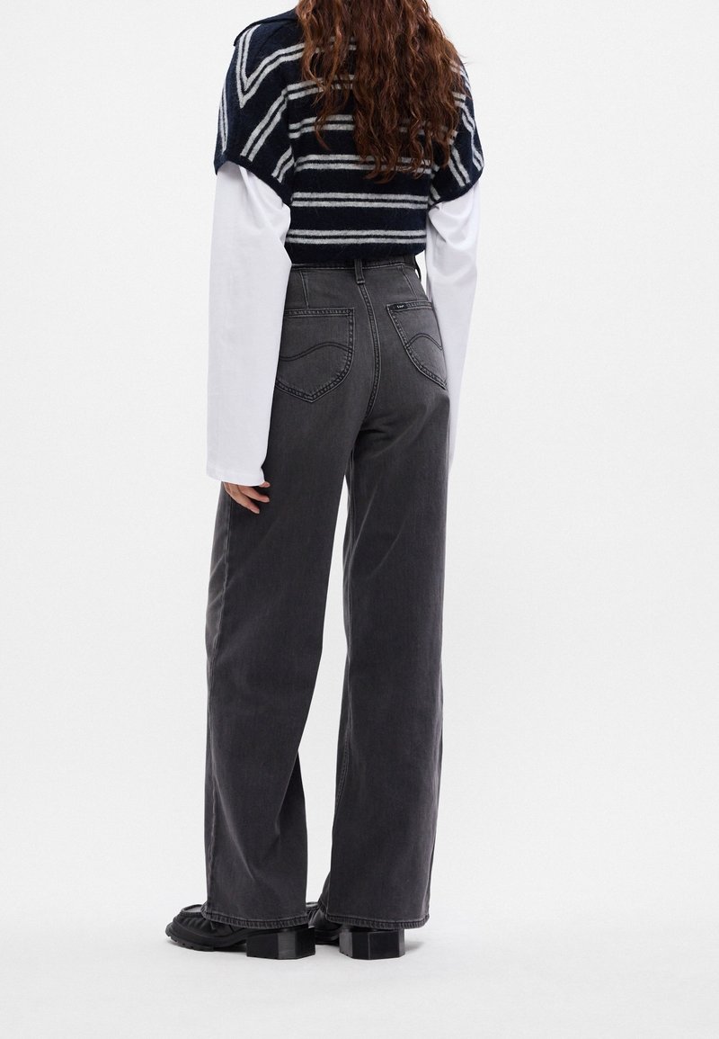 Femme aux longs cheveux portant un pull rayé noir et blanc par-dessus une chemise blanche, un jean ample foncé et des chaussures noires, debout de dos.