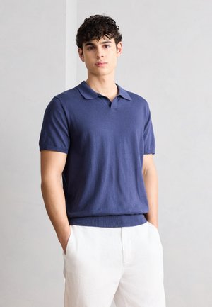 Fiatal férfi kék rövid ujjú kötött poloingben és fehér nadrágban, kezét a zsebében tartva egy sima fal előtt áll.