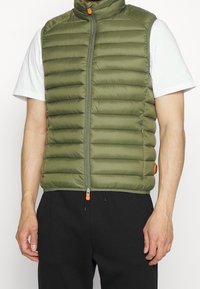 Chaleco de plumas verde oliva con cuello alto, costuras horizontales y un tirador de cremallera naranja. Lleva puesto sobre una camiseta blanca.