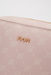 Ljust rosa texturerad handväska med ett subtilt mönster och guldtonat logo "JOOP!" präglat på ytan, med ett dragkedjestängning längs toppen.