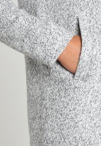 Cappotto di lana grigia con una sottile trama a macchie, dotato di una tasca laterale e un design pulito e sartoriale.