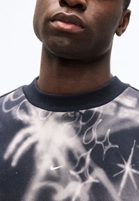 Κοντινό πλάνο του κάτω μέρους του προσώπου και του λαιμού ενός ατόμου που φοράει σκούρο φούτερ Nike με σχέδια λευκών αστεριών και στροβιλισμών.