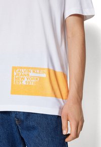 Biały bawełniany t-shirt z pomarańczowym nadrukiem graficznym przy dole, na którym widnieje napis "CALVIN KLEIN JEANS NEW YORK EST. 1978" w pogrubionych literach.