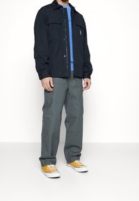 Black zip-up jacket over a blue shirt, paired with gray trousers and yellow sneakers. Plain design with buttons and pockets on the jacket.