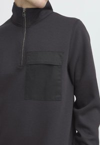 Hanorac negru cu fermoar și guler înalt, având o buzunare frontală dintr-un material de contrast. Textură moale, design minimalistic.